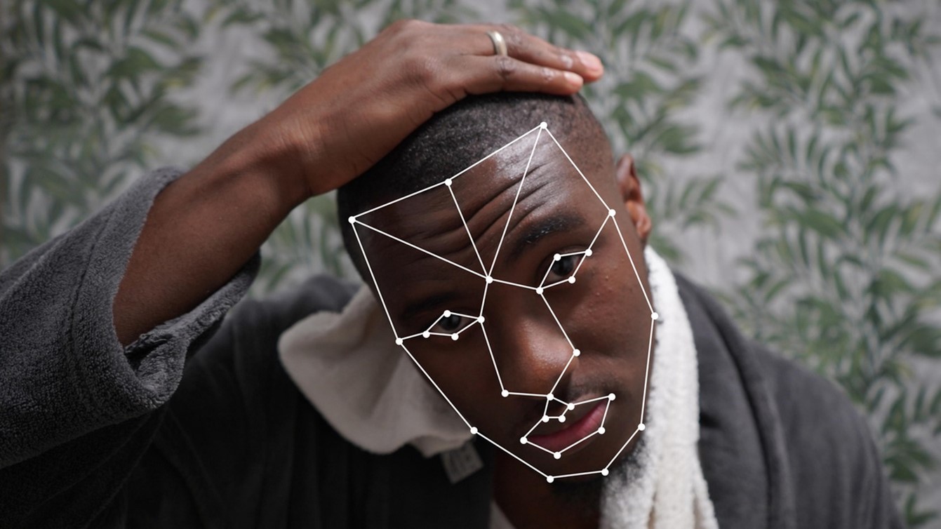 A young black man looks into the camera. He is wearing a dressing gown and has a towel around his neck, and there is green wallpaper behind. His face is mapped with a white wireframe.