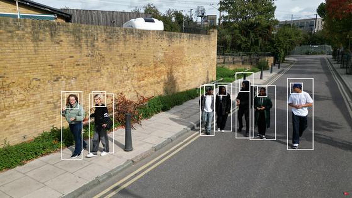 Young adults are walking on a street. Two young women are on the pavement to the left of the image and five young men walk in the road. The road is empty of traffic and the people in the picture are relaxed and appear to be engaged in chatting with each other.  Overlaid on the image are identification boxes. All of the figures have ID boxes around them. 